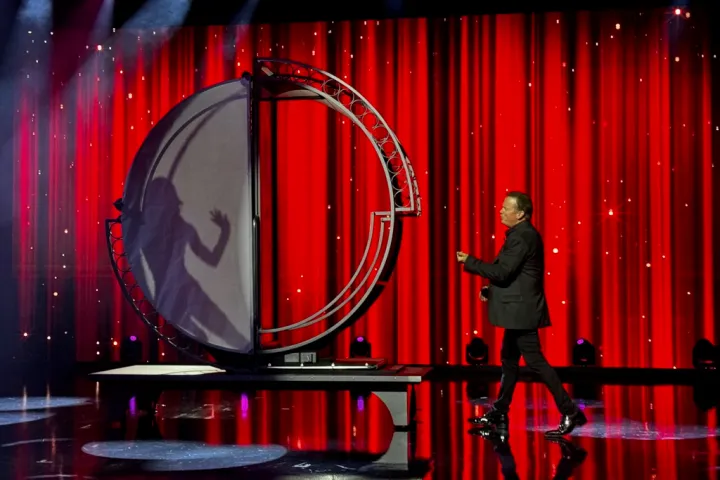 Magician on stage with a spinning illusion and shadow of a woman on red curtain background.