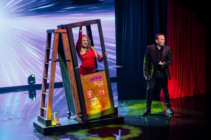 Magician on stage with assistant in red dress inside a wooden frame, colorful lights and curtains in background.