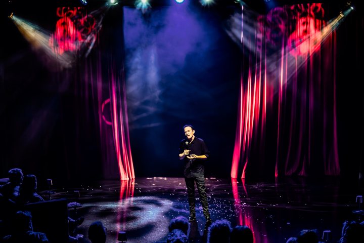 Performer on stage with spotlight, red curtains, and audience in shadow.