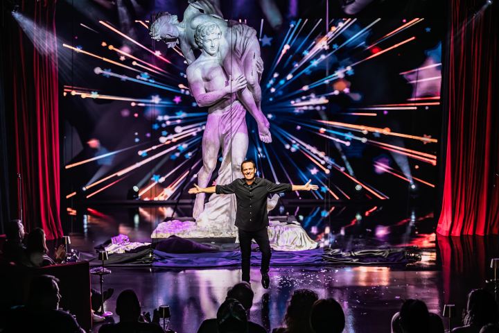 Performer on stage with dynamic backdrop featuring classical statue and colorful light effects.