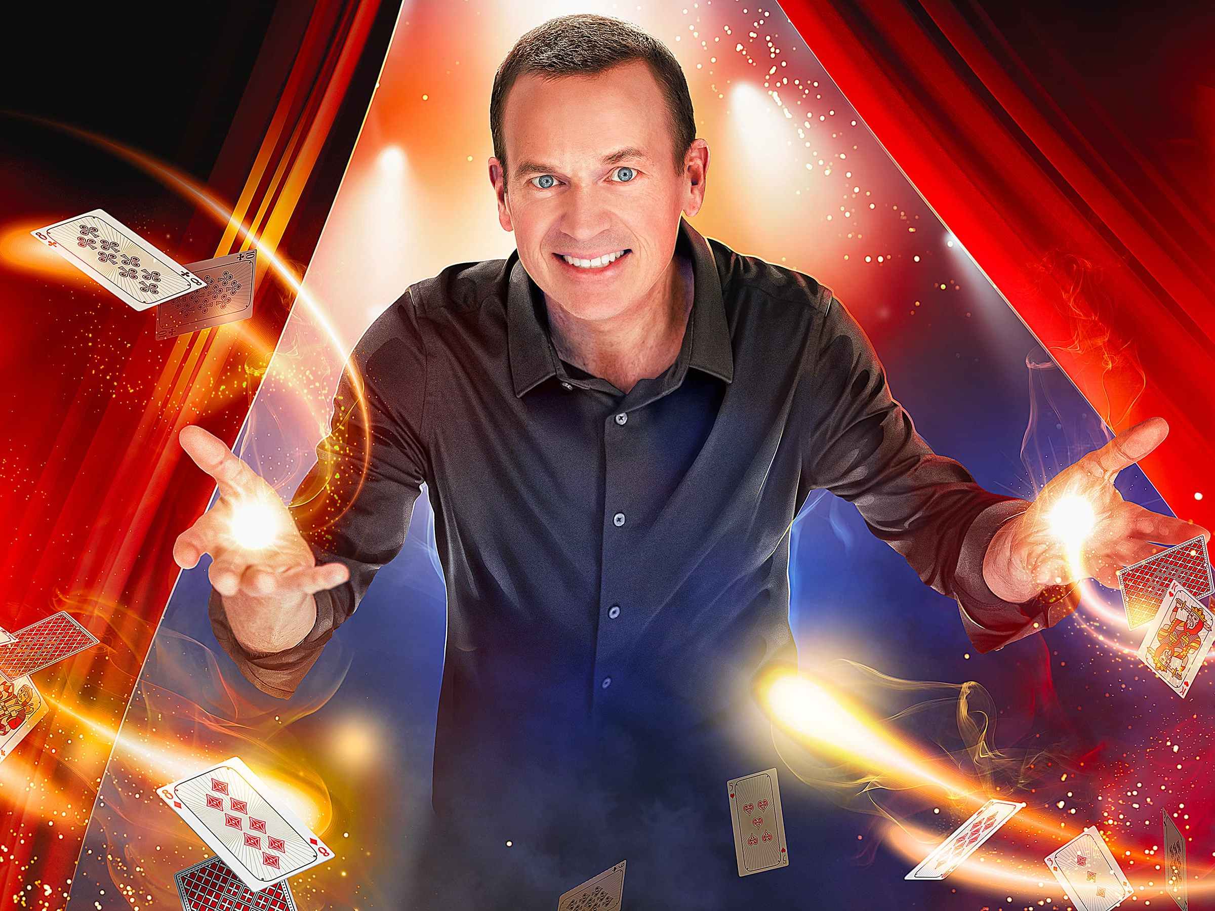 Magician on stage with playing cards and fiery lights, red curtains in background.